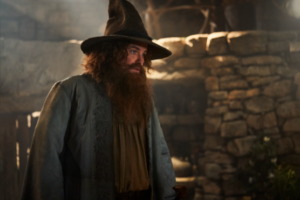 Rory Kinnear as Tom Bombadil in The Rings Of Power, photographed from the waist up, standing in a dimly-lit room of his humble house. He has long curly reddish-brown hair and a beard. He wears a tall gray feathered hat, and a blue jacket over a white tunic with a brown belt.