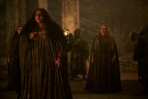 Sophia Nomvete as Disa in The Rings Of Power, standing in an underground stone hall, amongst a group of Dwarven women. She has long dark curly hair, and wears a floor-length gray gown. She has gold bracelets on her wrists and gold-dust on her fingers.