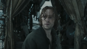 Charlie Vickers as Halbrand, standing at the back of a covered wagon, looking around. He has shoulder-length, shaggy brown hair, and wears brown rags.