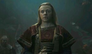 Jack Lowden as Sauron in The Rings Of Power. He has long blond hair, slicked-back, and wears black armor over a red-and-gold robe.