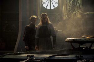 Charles Edwards as Celebrimbor and Charlie Vickers as Annatar, standing in a forge filled with smoke. Celebrimbor has short curly brown hair and wears a red robe with gold embroidery. Annatar has long blonde hair held back by a golden circlet and wears a brown leather apron over a white robe.