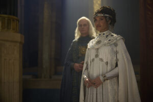 Cynthia Addai-Robinson as Miriel, standing with her hands clasped at her waist. She has dark hair hanging in loose ringlets, held back by a silver diadem encrusted with large dark gems. She wears a white gown with a black-and-white mosaic collar.