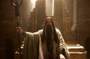 Ciaran Hinds as the Dark Wizard, seated on a stone throne carved with runes and hieroglyphs, in a cave between basalt pillars. He has long, straight dark brown hair, a long beard going gray, and bushy eyebrows. He wears white robes with a silver breastplate and gauntlets on both his wrists, and carries a horned staff in his right hand.