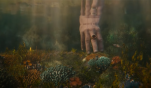 Underwater shot of a hand, reaching for the bottom of a shallow pool teeming with small fish and a few brightly-colored coral. The hand wears a single golden ring, in which a dark red gemstone is set.