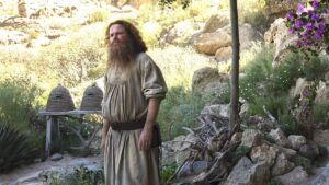 Rory Kinnear as Tom Bombadil, standing outside in a rock garden. Bee-hives sit on a wooden bench behind him, and a pile of branches. Tom has long curly reddish-brown hair and a bushy beard, and wears a white tunic with rolled-up sleeves and a brown leather belt.