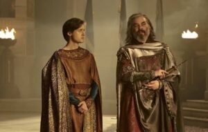 Leon Wadham as Kemen and Trystan Gravelle as Ar-Pharazon, standing side-by-side in a vast hall, talking. Kemen has short brown hair and wears a russet-brown robe with a gold cape and dark blue sleeves. Pharazon has shoulder-length curly gray hair and a beard, and wears a silver toga-like garment over a dark red robe, with a golden scepter in his hand.