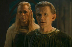 Charles Edwards as Celebrimbor, in close-up, with Charlie Vickers as Annatar standing behind him, looming over his right shoulder. Celebrimbor has short brown hair and wears a forest-green pleated velvet robe with holly leaves embroidered around the frilly collar. Annatar has long blonde hair held back by a golden circlet, and wears a brown leather apron over a plain white robe with moderate silver embroidery.