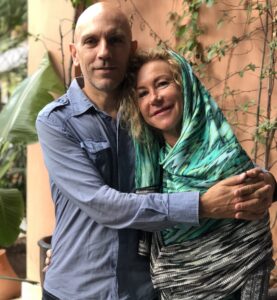 Luca Mosca standing with his arms wrapped around his sister Erica. Luca is wearing a long-sleeved blue shirt. Erica is wearing a blue-green headscarf and black-and-white dress.