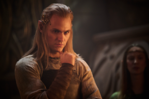 Charlie Vickers as Annatar in The Rings Of Power, standing with his right hand raised to his chin in a contemplative gesture. He has long blond hair held back by a serpentine gold circlet, and wears a brown leather apron over a silver robe with minimal silver embroidery around the collar. He also wears brown leather arm-wraps.