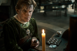 Charles Edwards as Celebrimbor in The Rings Of Power, seated at a small desk watching a candle burning. He has tousled brown hair streaked with soot and grime, and wears a brown leather apron over a dark green robe with holly leaves embroidered in spring green and gold around the collar. The tools and accoutrements of a jewelsmith are scattered about him.