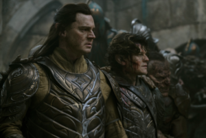 Benjamin Walker as Gil-galad and Robert Aramayo as Elrond, standing side-by-side, surrounded by Orcs. Gil-galad has long dark brown hair and wears a burnished gold breastplate emblazoned with the intertwining gold and silver branches of the Two Trees of Valinor over a long-sleeved pale yellow tunic with large pauldrons and vambraces. Elrond has short, tousled dark brown hair and wears a very similar suit of armor but in silver. He has a deep wound in his right cheek, and blood is running down his face.