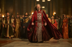 Trystan Gravelle as Pharazon in The Rings Of Power, striding towards the camera with his hands by his side, as a wind whips at him. He has long dark hair going gray and a beard streaked with black. He wears a crimson robe with a long cloak and an ornamental silver breastplate with numerous belts around his waist.