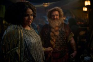 Sophia Nomvete as Disa and Owain Arthur as Durin IV, in a crowded market. Disa has curly dark hair piled up on her head, and wears a silver-and-gold mantle over a pleated gray dress encrusted with gold. Durin has bushy reddish-brown hair and a long braided beard, and wears a rust-red studded breastplate over a red-and-gold tunic with red leather armbands.