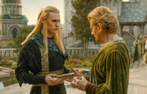 Charlie Vickers as Sauron and Charles Edwards as Celebrimbor, standing outdoors in the main square of Eregion. Sauron has long blonde hair and wears a black robe with gold trim and a wide gold belt. He is presenting a gilded hammer to Celebrimbor. Celebrimbor has short brown hair and wears a green velvet gown.