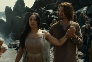 Cynthia Addai-Robinson as Miriel with Lloyd Owen as Elendil standing behind her, holding her arm steady as she walks up a rocky path between rows of soldiers and citizens. Miriel has long dark curly hair and wears a sleeveless white gown. Elendil has shaggy shoulder-length brown hair and wears a brown tunic.