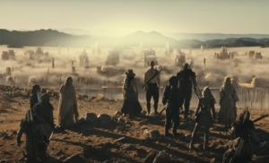 Josha Stradowski as Rand al'Thor and Rosamund Pike as Moiraine Damodred in The Wheel Of Time stand, accompanied by a group of Aiel warriors and at least one Wise One, at the edge of a cliff, looking out over a barren valley filled with fog and the ruins of ancient skyscrapers encircled by a tall wall. Rand has short reddish-brown hair and wears a white shirt and brown trousers, with a sword strapped to his back. Moiraine wears a hat, and a long dress. The Aiel warriors carry spears and wear lightweight sand-brown leather armor, while the Wise One is swathed head-to-toe in white fabric.