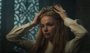 Ceara Coveney as Elayne Trakand in The Wheel Of Time, lowering a golden crown encrusted with rubies and pearls onto her head. She has long golden hair and wears a long-sleeved white dress.