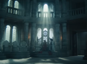 Wide shot of the Hall of Sitters in the White Tower, from The Wheel Of Time. A woman in a long red gown is sitting on the Amyrlin Seat, with a dark-haired woman in a white dress sitting on the armrest at her left hand.