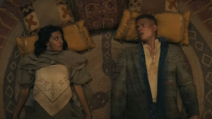 Madeleine Madden as Egwene al'Vere and Josha Stradowski as Rand al'Thor, lying on their backs side-by-side on the floor of a tent, with pillows and rugs underneath them. Egwene has short dark hair and wears a gray top with baggy sleeves over a white tunic dress. Rand has short reddish-brown hair and wears a sand-brown coat over a partially unbuttoned off-white shirt.