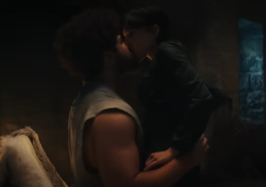 Marcus Rutherford as Perrin Aybara, seated on a bed, with Isabella Bucceri as Faile on his lap, kissing him. Perrin has frizzy dark hair and a beard, and wears a sleeveless white shirt. Faile has short dark hair and wears a long-sleeved black jacket.