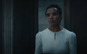 Zoe Robins as Nynaeve al'Meara. She has long dark hair pulled back in a braid. She's wearing a long-sleeved white dress with a narrow oval cutout in the front.