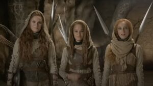 Ragga Ragnars as Bain, Ayoola Smart as Aviendha, and Maja Simonsen as Chiad standing in a row in a dimly-lit room with stone walls. All three have long reddish-brown hair and wear lightweight brown leather armor over sand-brown garments with hoods and black scarves. They have spears strapped to their backs and small bucklers at their sides.