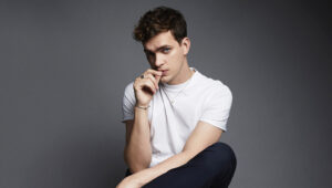 Luke Fetherston, sitting in front of a gray background, wearing a white t-shirt and black pants. He has short reddish-brown hair.