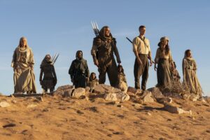 (From left to right) Nukâka Coster-Waldau as Bair, Synnøve Macody Lund as Melindhra, Daniel Henney as Lan Mandragoran, Björn Landberg as Rhuarc, Josha Stradowski as Rand al'Thor, and Rosamund Pike as Moiraine Damodred, standing on a rugged hilltop in a desert under a cloudless sky. Bair has long white hair and wears a white dress with a hood and shawl. Melindhra has blonde hair under a brown hood, and wears lightweight brown leather armor, a skirt of tassels, and brown pants, with three short spears strapped to her back. Lan wears a gray hood and baggy gray tunic. Rhuarc has red hair under a sand-brown hood, with brown leather armor and trousers made from a coarse brown fabric. Rand has short curly reddish-brown hair and wears a white shirt with rolled-up sleeves, dark trousers, and a sword strapped to his back. Moiraine has long brown hair under a large hat, and wears a brown shawl over a sand-brown top, with a long gray dress.