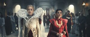 Olivia Williams as Morgase Trakand and Shohreh Aghdashloo as Elaida, walking side-by-side down a corridor in the White Tower, between rows of soldiers in red-and-gold uniforms and Novices in white dresses. Morgase has short blonde hair under a gold tiara encrusted with rubies. She wears a large white heart-shaped lace collar, and a long-sleeved white lace coat over a long dark red gown. Elaida has short jet-black hair. She wears a crimson coat over a dark red gown with a heavy gold necklace, large gold earrings, gold bracelets on both arms, and multiple gold rings.