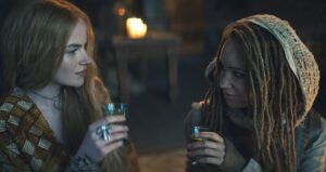 Ceara Coveney as Elayne Trakand and Ayoola Smart as Aviendha, drinking together and smiling. Elayne has long reddish-brown hair and wears a gold dress. Aviendha has long reddish-brown hair in locs, and wears a sand-brown hood made from a coarse fabric, with lightweight brown leather armor and fingerless gloves.