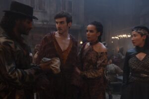 Donal Finn as Mat Cauthon, Zoe Robins as Nynaeve al'Meara, and Kae Alexander as Min Farshaw in The Wheel Of Time, standing in a smoky room amongst a crowd of colorfully costumed revelers, speaking to a man in a black hat who is handing them something wrapped in a cloth. Mat has short dark brown curly hair and a patchy beard. He wears an oversized frilly maroon coat over a low-collared saffron-yellow shirt. Nynaeve, standing beside him with her hand on his arm, has long dark hair in a braid and dark eye makeup. She wears a red, black and gold long-sleeved dress with shoulder cutouts. Min has black hair in a slicked-back mullet, with a black-and-white headwrap. She wears a black leather bolero over a black-and-gold top, and a gray dress with pockets. She has a throwing knife in a small scabbard on the front of her belt.