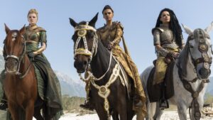 (from left to right), Ceara Coveney as Elayne Trakand, Madeleine Madden as Egwene al'Vere, and Zoe Robins as Nynaeve al'Meara, on horseback. Elayne has blonde hair in a bun, and wears a gauzy green dress with gold armor. Egwene has short dark hair under a gold tiara and wears gold armor over a long gold gown with a serpentine flame pattern. Nynaeve has long dark hair, and wears a padded silver vest and armor over a yellow dress.