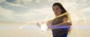 Rosamund Pike as Moiraine Damodred, standing in a desert, channeling golden threads of the One Power into a glowing orb she's holding out in front of her. She has long dark brown hair, and a blue gemstone on a slender gold diadem sits upon her brow. She wears an intricate gold lattice-work chest-piece over a sleeveless blue dress.