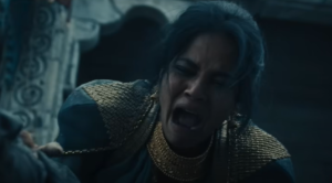 Priyanka Bose as Alanna Mosvani, weeping over someone's body. She has long dark hair covered in ash. She's wearing a green dress with gold epaulets and a gold collar.