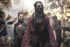 Donal Finn as Mat Cauthon, Kae Alexander as Min Farshaw, and Ceara Coveney as Elayne Trakand, walking down a busy street in Tanchico. Mat has short curly dark brown hair under a long black lace veil adorned with flower appliques. He wears an oversized semi-transparent frilly maroon coat over a low-collared saffron-yellow shirt with high-waisted brown floral-patterned trousers. Min has jet-black hair in a slicked-back mullet, with a camouflage headwrap and half-veil covering the lower half of her face. She wears a black leather bolero over a black-and-gold top with a long gray dress, and carries a throwing knife in a scabbard on her belt. Elayne has long reddish-gold hair under a white turban with a gauzy white half-veil covering the lower half of her face, and wears a leopard-patterned jacket.