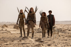 (Left to right) Maja Simonsen as Chiad, Ragga Ragnars as Bain, Ayoola Smart as Aviendha, and Marcus Rutherford as Perrin Aybara, walking through a desert. Aviendha, Bain and Chiad all have long reddish-brown hair. They wears very similar outfits: sand-brown hoods, black scarves, brown leather armor over sand-brown tunics, skirts and trousers, with short spears strapped to their backs. Perrin has short dark frizzy hair and a beard, and wears a dark green woven-leather vest over a faded red long-sleeved shirt with olive-green trousers and brown boots.