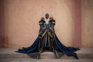 Daniel Francis as High Lord Turak, standing in front of a pink stucco wall. He is bald. He wears an intricate collar of bone over a black feathered mantle, over a black-and-gold scale-patterned robe with a long dark blue train. The first two fingernails on both his hands are extremely long and bladed.