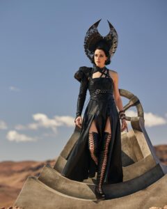 Natasha O’Keeffe as Lanfear, descending from a stone dais in the middle of the desert, upon which stands a circular throne. She has short jet-black hair under a black crescent-moon headdress. She wears a black leather harness over a black dress wit thigh-high slits and one sleeve covering her right arm, with thigh-high black lace-up high-heeled boots.